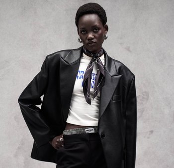 Donna nera con blazer oversize in pelle nera, t-shirt corta bianca con grafica, pantaloni a quadri, sciarpa nera annodata al collo e orecchini a cerchio.