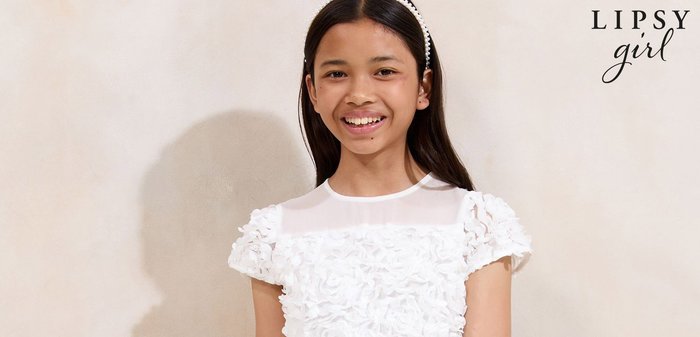 Jeune fille souriante, vêtue d'une tenue blanche avec des détails floraux en 3D et un panneau supérieur en voile, debout devant un fond neutre.