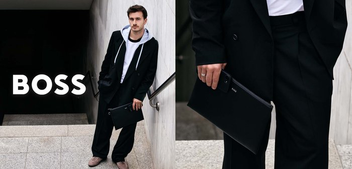 Hombre con abrigo negro y ropa informal está en las escaleras sosteniendo una bolsa negra con cremallera y un smartphone, el logo de BOSS es visible en el lado izquierdo.