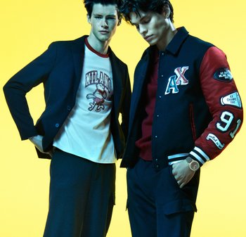 Czarna marynarka, biała koszulka z czerwoną lamówką i grafiką, czerwono-granatowa kurtka varsity z naszywkami, ciemne spodnie i zegarek, na żółtym tle.