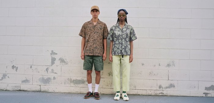 Deux personnes se tiennent côte à côte devant un mur de briques blanches, portant toutes deux des chemises à motifs et des vêtements décontractés, face à l'avant avec des expressions neutres.