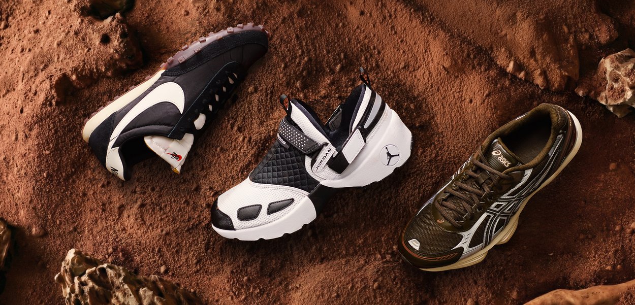 Drie sportschoenen—zwarte Nike, witte en zwarte Air Jordan, en bruine Asics—op een roodbruine rotsachtige ondergrond gelegd.