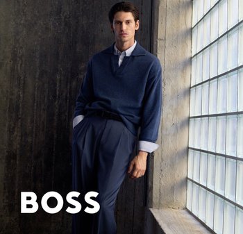 Man draagt een marineblauwe trui en broek, leunt tegen een muur nabij een groot raam van glazen blokken, met de tekst "BOSS" in de hoek.