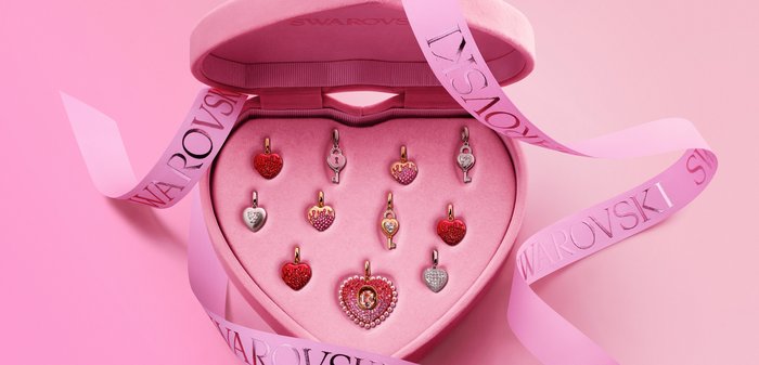 Hartvormige en sleutelvormige Swarovski-kristalbedels in een roze fluwelen hartendoos met een roze Swarovski-lint op een roze achtergrond.