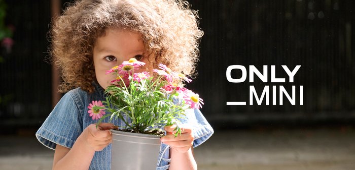 Barn med krøllet hår holder en potte med lyserøde margueritter og lugter til dem, tekst "ONLY _MINI" på en mørk baggrund ved siden af.