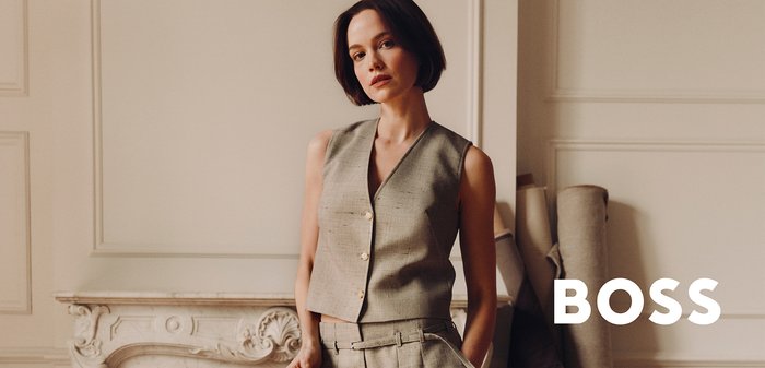 Woman with short dark hair wearing a beige sleeveless vest and matching trousers, holding a jacket, standing in an elegant room.