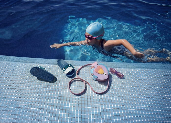 Enfant portant un bonnet de bain et des lunettes de natation nage dans la piscine près du bord avec des sandales, un sac en forme de poisson et des lunettes de soleil roses posés sur les carreaux.