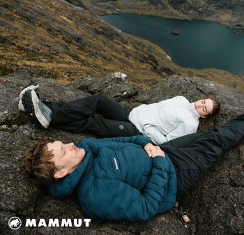 Mammut Daunenjacken in Dunkelblau und Weiß mit schwarzen Hosen, die auf felsigem Terrain nahe einem See ruhen und eine robuste Outdoor-Umgebung präsentieren.