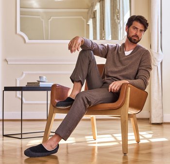 Brauner Strickpullover, graue maßgeschneiderte Hose und schwarze Slippers. Sitzt in einem Ledersessel mit Holzbeinen. Minimalistische Inneneinrichtung.
