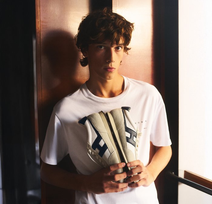 Giovane uomo con capelli ricci, indossa una maglietta bianca e tiene delle scarpe da ginnastica bianche e nere rivolte verso la macchina fotografica, su uno sfondo di legno e luminoso.