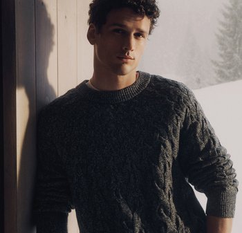 Ung man i mörk kabelstickad tröja som lutar sig mot en trävägg med ett snöigt landskap suddigt i bakgrunden.