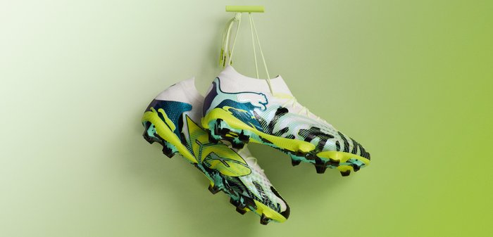 Chaussures de soccer Puma blanches et jaunes avec des accents bleus et noirs suspendues par les lacets sur un fond à dégradé vert.