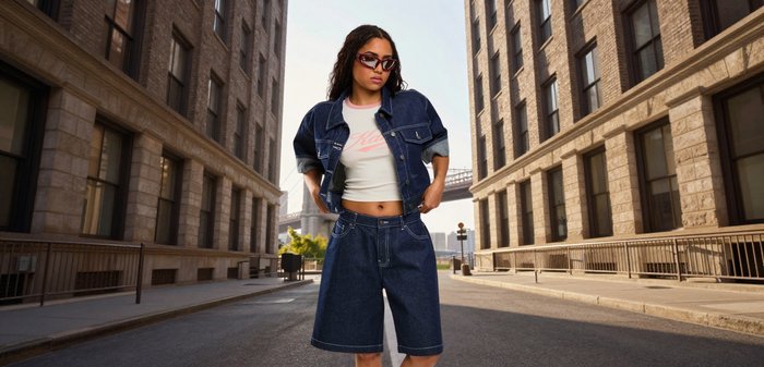 Frau trägt übergroße dunkle Jeansjacke und Shorts, weißes kurzes T-Shirt und Sonnenbrille, steht auf einer leeren Stadtstraße zwischen Gebäuden.