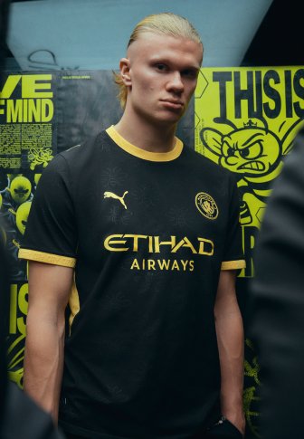 Junger Mann mit blondem Haar trägt ein schwarz-gelbes Manchester City Fußballtrikot und steht vor einem gelb-schwarzen Werbehintergrund.