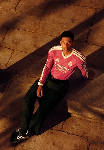 Junger Mann trägt ein pinkes Adidas Real Madrid Trikot und sitzt auf einem Steinboden mit Sonnenlicht und Schatten.
