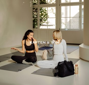 Dvě ženy sedí na šedých jógových podložkách v prostorné místnosti s velkými okny. Jedna má na sobě černý sportovní top a legíny; druhá má světle šedé oděvy pro odpočinek. Na podlaze je černá taška a průhledná lahev s vodou.