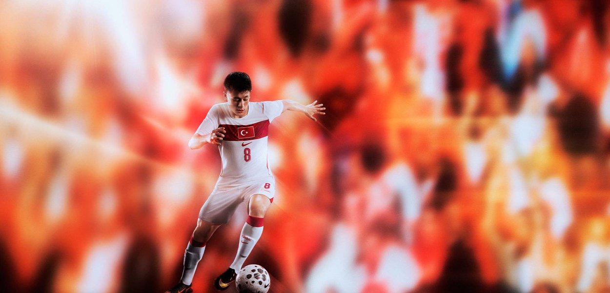 Joueur de football en tenue blanche et rouge de la Turquie contrôle le ballon sur le terrain avec une foule orange floue en arrière-plan.