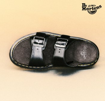 Zwarte leren Dr. Martens slippers met twee gespen en zichtbare gele stiksels langs de rand van de zool, op een beige achtergrond.