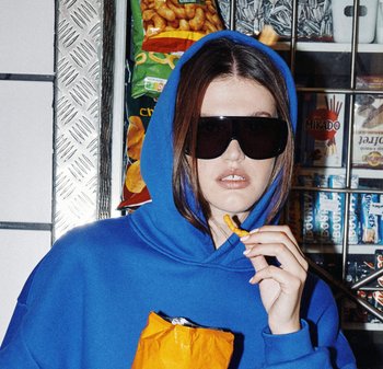 Niebieska bluza z kapturem, oversize'owe czarne okulary przeciwsłoneczne, trzyma przekąskę. W tle różne przekąski w opakowaniach na metalowych półkach.