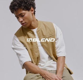 Jonge man met krullend haar, gekleed in een beige vest over een wit shirt met lange mouwen, zittend en omlaag kijkend tegen een effen achtergrond.