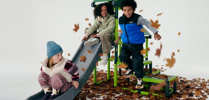 Des enfants jouent sur un toboggan gris entouré de feuilles d'automne. Parmi les éléments, on trouve un ensemble de jeux vert, des vêtements d'hiver colorés et des textures variées.