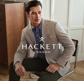 Homme portant un blazer beige, une chemise bleue et un pantalon vert foncé, assis sur une chaise dans une pièce avec des meubles en bois et des murs lambrissés.