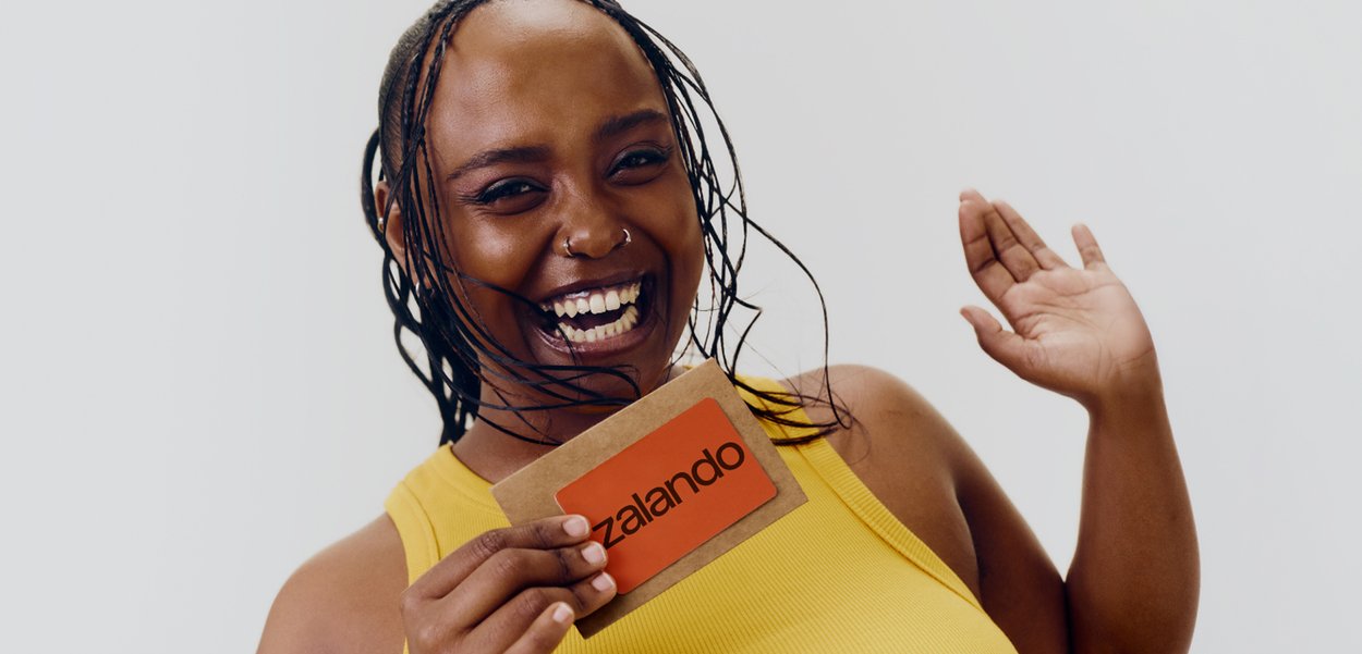 Donna sorridente con canottiera gialla che tiene una carta regalo arancione Zalando e saluta con l'altra mano su uno sfondo semplice.