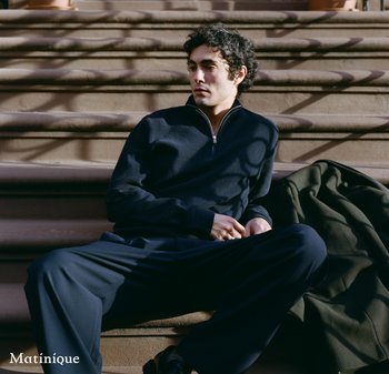 Young man in dark casual wear sits on outdoor stone steps with coat beside him, looking to his left in soft sunlight.