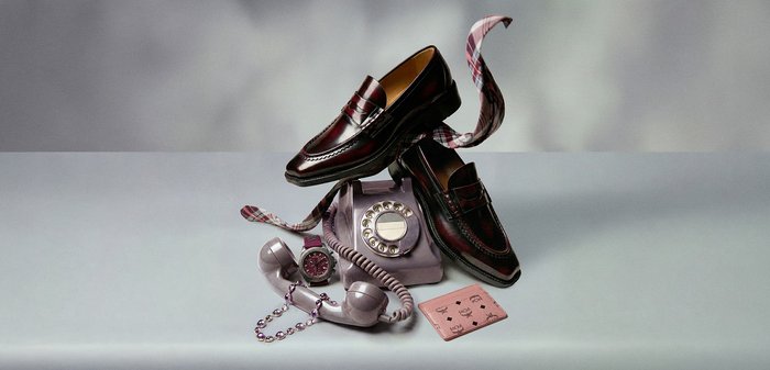 Pár lesklých burgundských mokasín odpočívajících na vintage fialkovém rotarovém telefonu spolu s kostkovanou šálou, ladícími hodinkami, korálkovým náramkem a růžovou peněženkou.