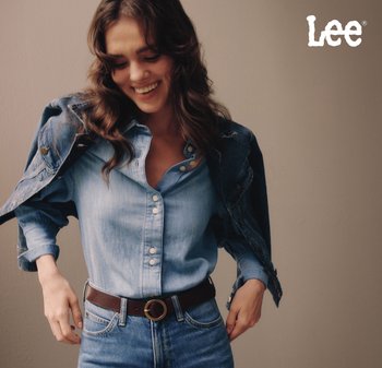 Chemise en denim bleu clair à boutons avec des manches retroussées, associée à une ceinture marron foncé avec une boucle circulaire, et une veste en denim jetée sur l'épaule.