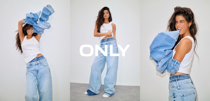 Femeie cu păr lung și închis la culoare, purtând un top alb fără mâneci, crop, și blugi largi, cu talie înaltă, de culoare albastru deschis, stând în picioare pe un fundal simplu.