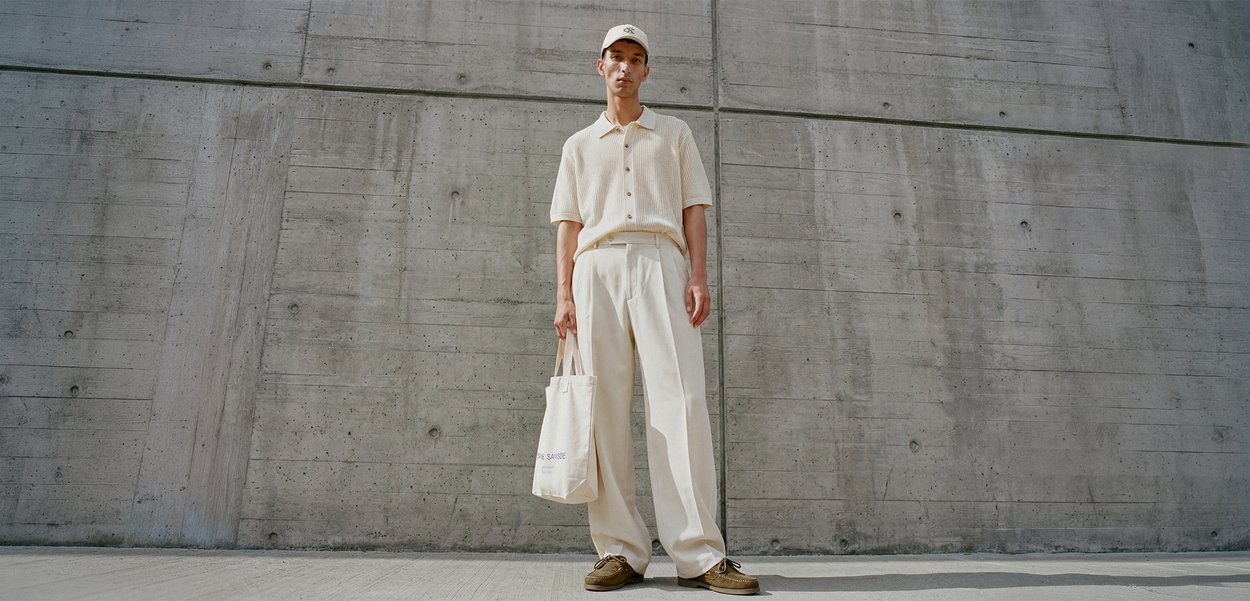 Joven con camisa beige, pantalones anchos y gorra, sosteniendo una bolsa tote blanca, de pie frente a una pared de hormigón al aire libre.