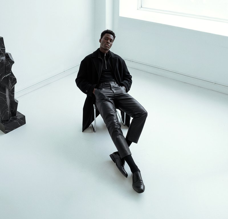 Black coat over a black turtleneck, teamed with black leather trousers and shoes. The background showcases a minimalist white space with a geometric black sculpture.