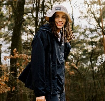 Schwarze leichte Jacke mit Reißverschluss, kombiniert mit einem beigen Bucket Hat. Umgeben von Bäumen in einer natürlichen Umgebung.