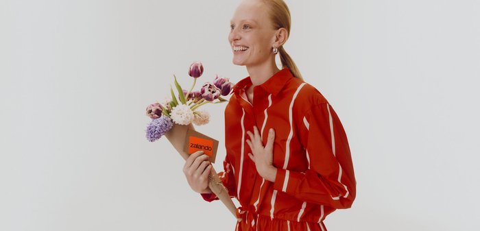 Femme en tenue rayée rouge souriante, tenant un bouquet de fleurs mixtes enveloppé dans du papier avec une étiquette Zalando.