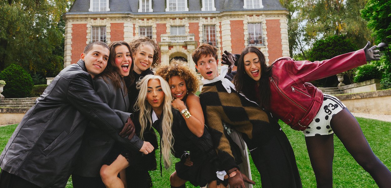 Sept jeunes adultes posant avec énergie, souriant et se penchant en avant sur une pelouse verte devant un bâtiment historique en briques rouges avec des arbres à proximité.