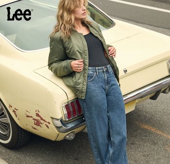 Giacca bomber verde oliva sopra una maglietta nera, abbinata a jeans blu regular fit. Sullo sfondo si vede un'auto gialla vintage con una finitura graffiata.