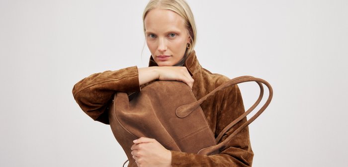 Bruine suede schoudertas met twee lange handvatten, met een minimalistisch ontwerp en een zachte textuur, vastgehouden door een persoon in een bijpassende bruine jas.