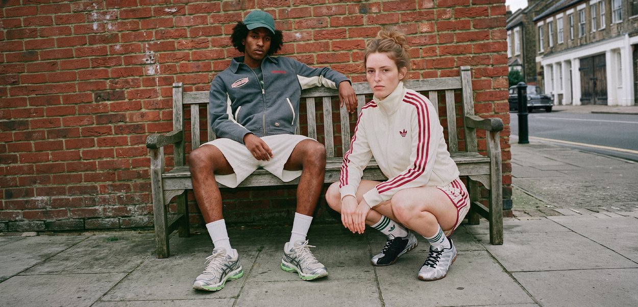 Deux jeunes en tenue sportive, l’un assis sur un banc en bois et l’autre accroupi à côté, devant un mur en briques rouges en extérieur.