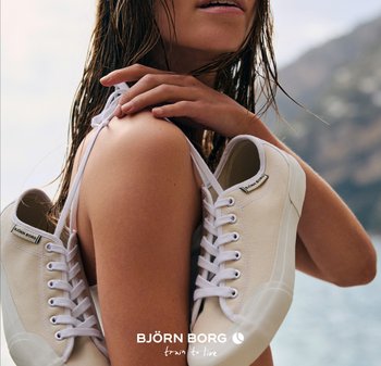 Kvinna med blött hår som håller vita Björn Borg-sneakers över axeln vid vatten och berg i bakgrunden.