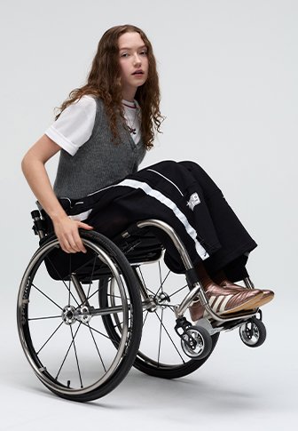 Persona su una sedia a rotelle che indossa un gilet grigio sopra una camicia bianca e pantaloni neri con strisce bianche. La sedia a rotelle presenta accenti metallici e ruote grandi.