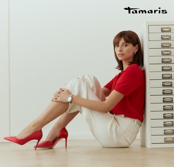 Woman in red top, beige pants, and red high heels sitting on floor, leaning against a white drawer unit, neutral background.