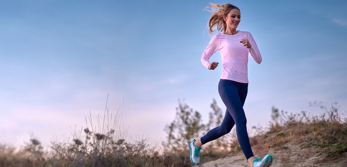 Lichtroze atletisch shirt met lange mouwen, marineblauwe legging en groene hardloopschoenen. Hardloper op een onverharde weg met gras en bomen op de achtergrond.