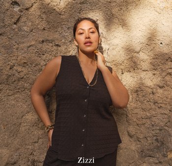 Selvsikker kvinde iført sort ærmeløs knapbluse og guldsmykker, stående foran en tekstureret, soloplyst beige væg.