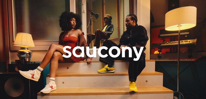 Saucony sneakers: white with red accents and grey sole; yellow sneakers with blue accents. Background features soft lighting and wooden stairs.
