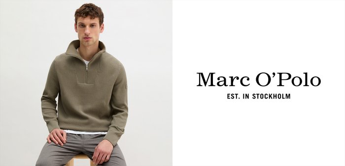 Mężczyzna w beżowym swetrze z półzamkiem i prążkowanym wzorem oraz szarych spodniach siedzi na drewnianym stołku na tle gładkiej ściany.