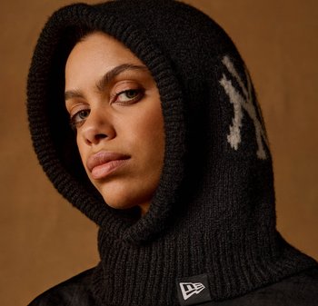 Person iført en sort strikket hættehalstørklæde med et hvidt "NY"-mønster, der kigger let til siden mod en brun baggrund.