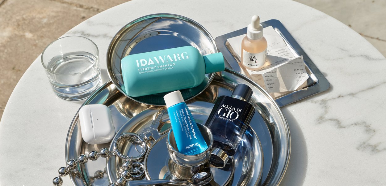 Bottiglia di shampoo blu, tubi per la cura della pelle blu, profumo nero, bicchiere d'acqua, custodia bianca per auricolari e bottiglia con contagocce su vassoi rotondi d'argento.