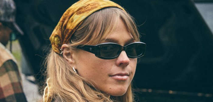 Jonge vrouw met zwarte zonnebril en een geel patroon hoofddoek, met lang blond haar en een grijze jas, buiten bij apparatuur.