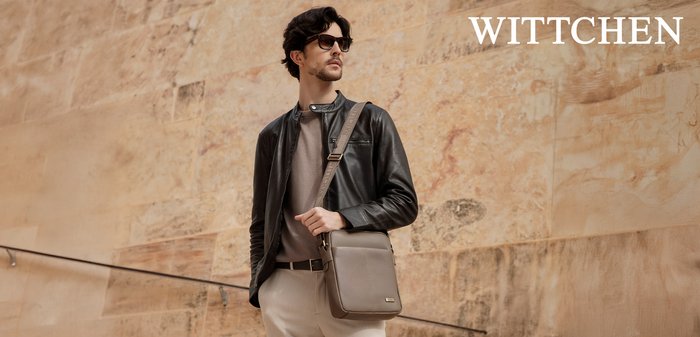 Mann mit schwarzer Lederjacke und Sonnenbrille trägt graue Umhängetasche und steht vor einer beigen Steinmauer mit WITTCHEN-Logo.